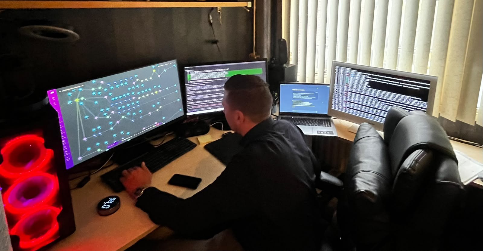 De oprichter van HV Infra aan het werk achter een opstelling met meerdere schermen, met een netwerkvisualisatie en code in beeld